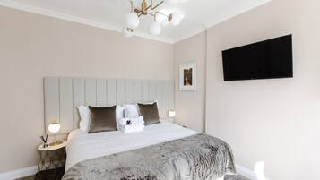 A neatly made bed with a grey blanket, a wall-mounted TV, and a modern chandelier.