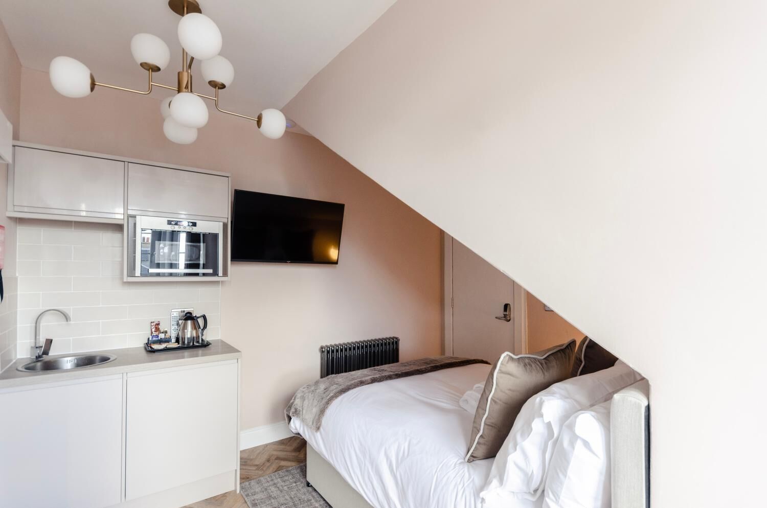 A compact bedroom with a bed, a TV, a kitchenette, and a radiator.
