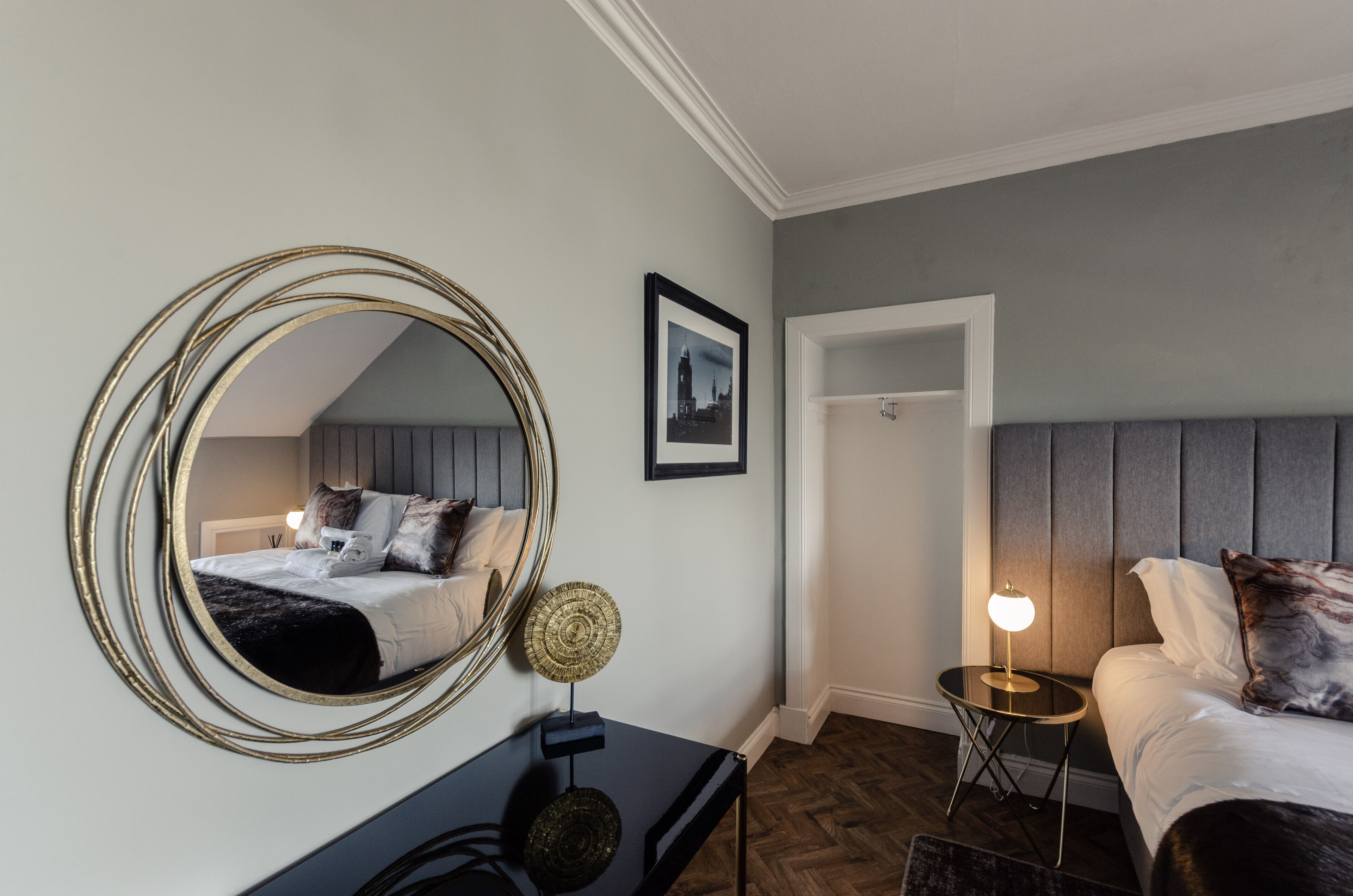 A hotel room with a bed, a round mirror, a small table with a lamp, and a framed picture on the wall.