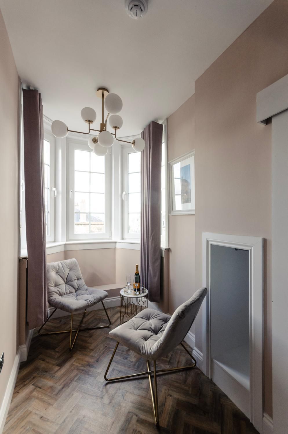 A modern room with a herringbone wood floor, two grey upholstered chairs, a small round table with a bottle and glass, a pendant light, and a large window with curtains.