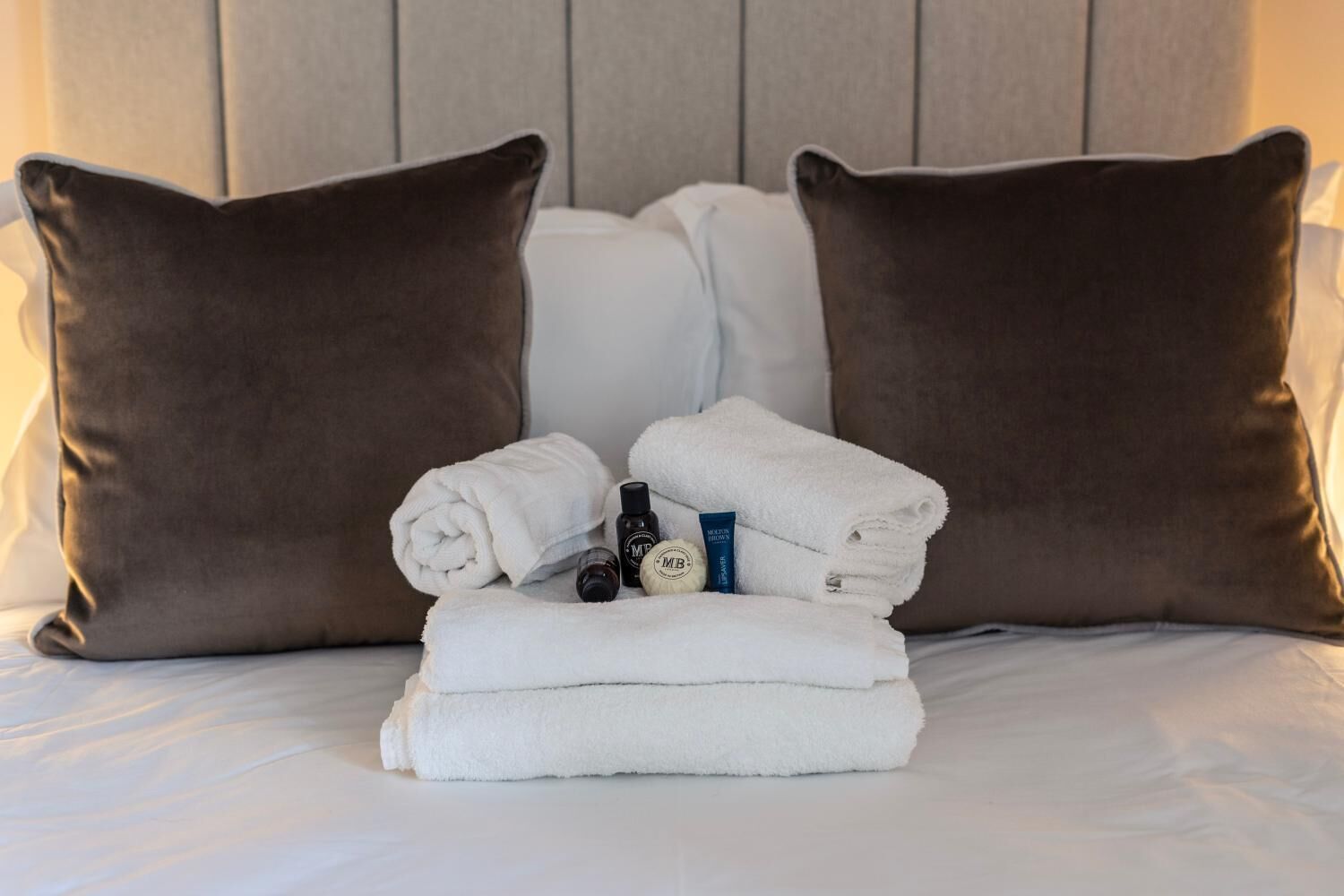Una cama bien hecha con dos almohadas marrones, una pila de toallas blancas y una botella de loción.