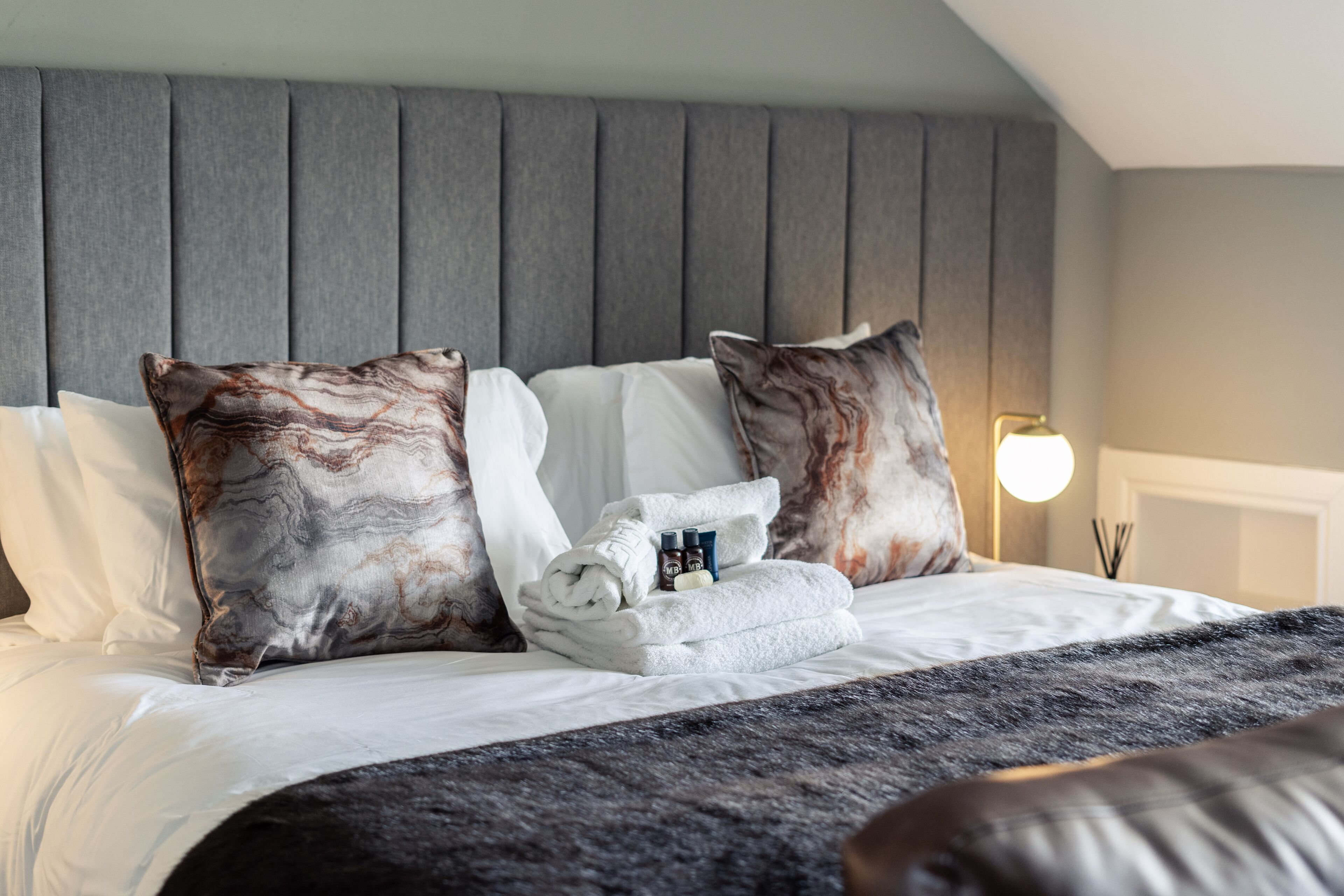 A neatly made bed with a grey headboard, white pillows, and a folded towel with toiletries.