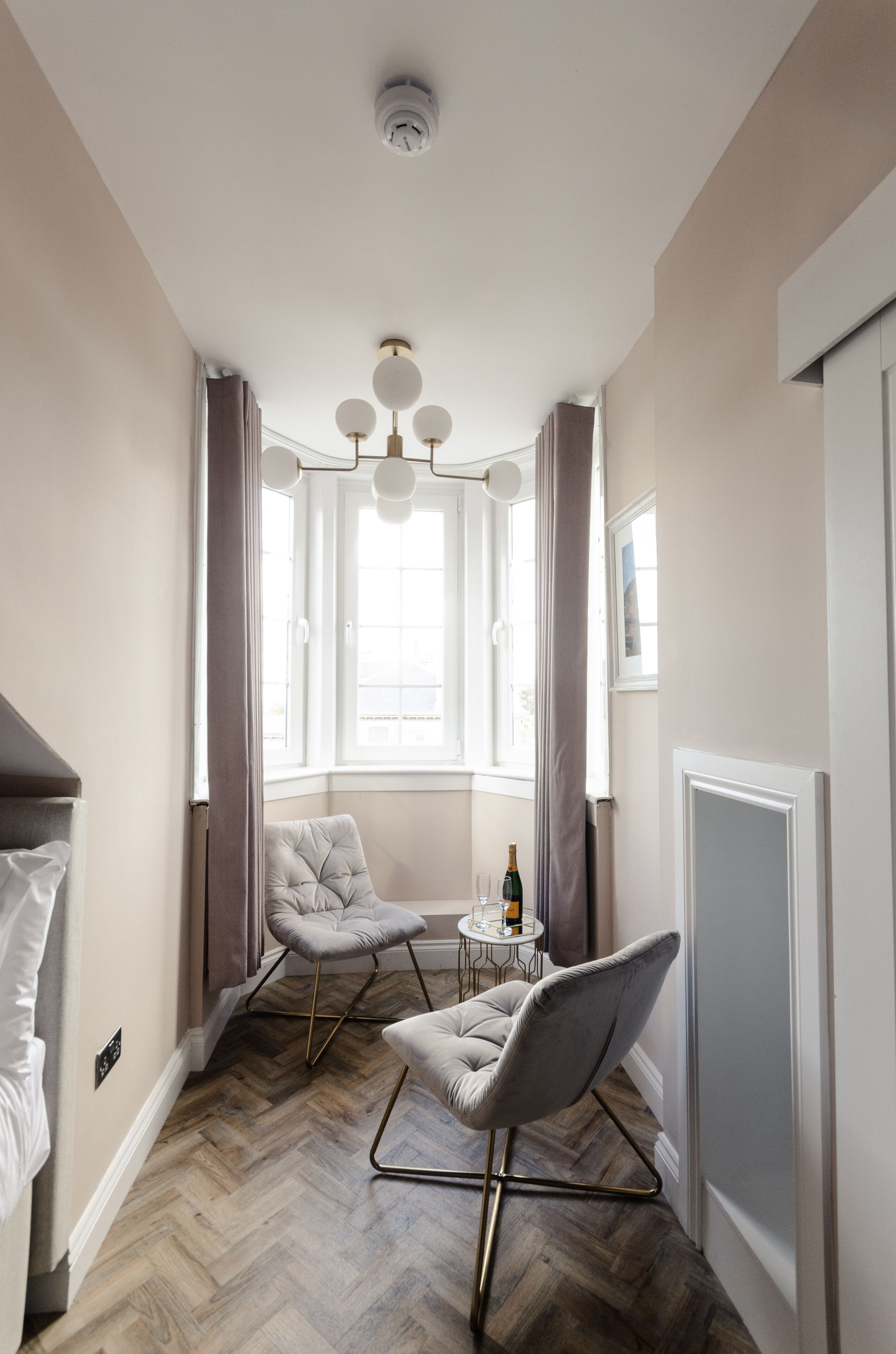 A modern room with a herringbone floor, a tufted armchair, a round table with a bottle, and a window with curtains.