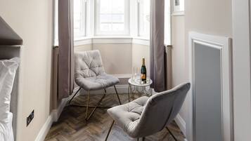 Une pièce moderne avec un parquet à chevrons, un fauteuil capitonné, une table ronde avec une bouteille, et une fenêtre avec des rideaux.