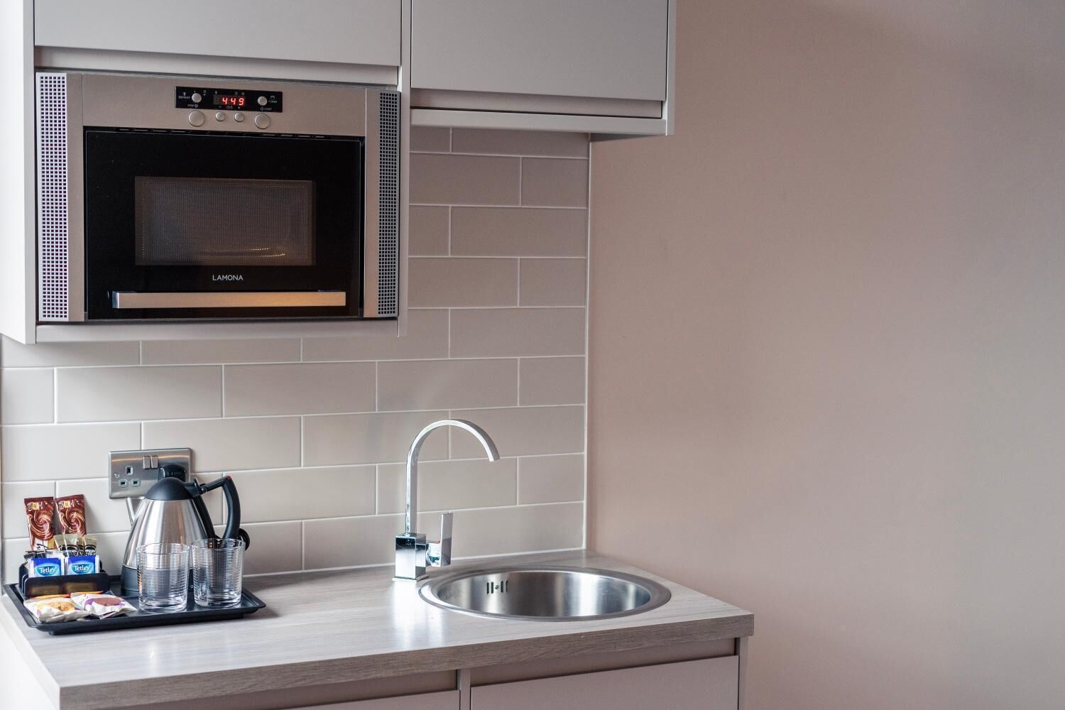 Una cocina moderna con microondas integrado, pileta de acero inoxidable y tetera sobre la encimera.