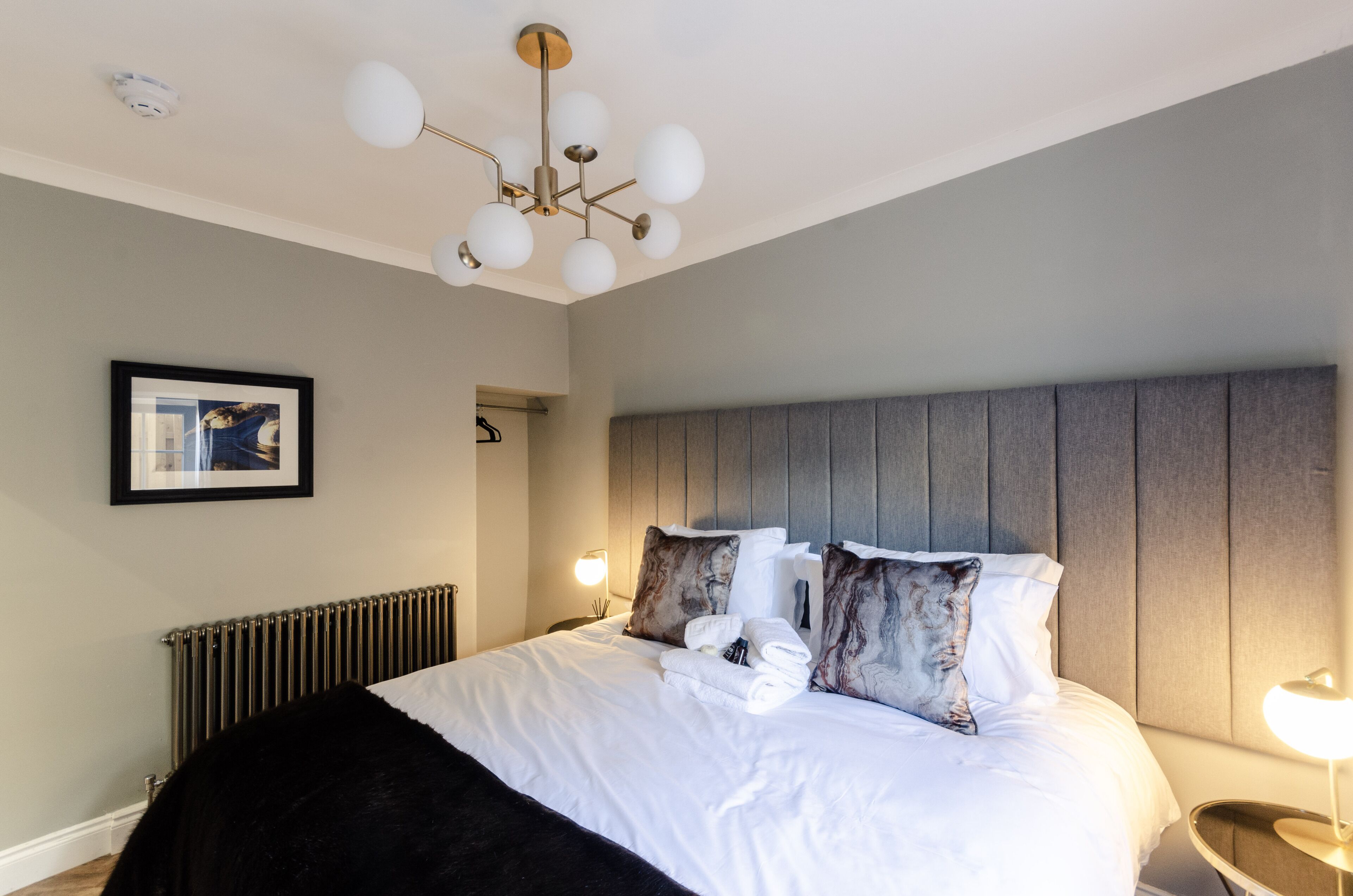 A neatly made bed with white linens, a grey headboard, and a modern chandelier.