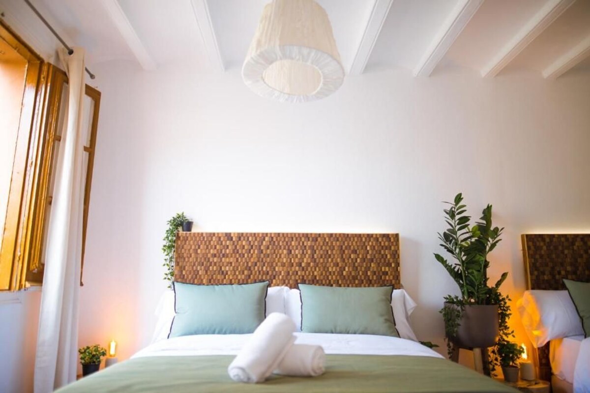 Ein ordentlich bezogenes Bett mit einer grünen Tagesdecke, zwei Kissen und einem zusammengerollten Handtuch. Am hölzernen Kopfteil ist ein gewebtes Muster zu sehen, und auf beiden Seiten des Bettes stehen je ein Blumentopf.