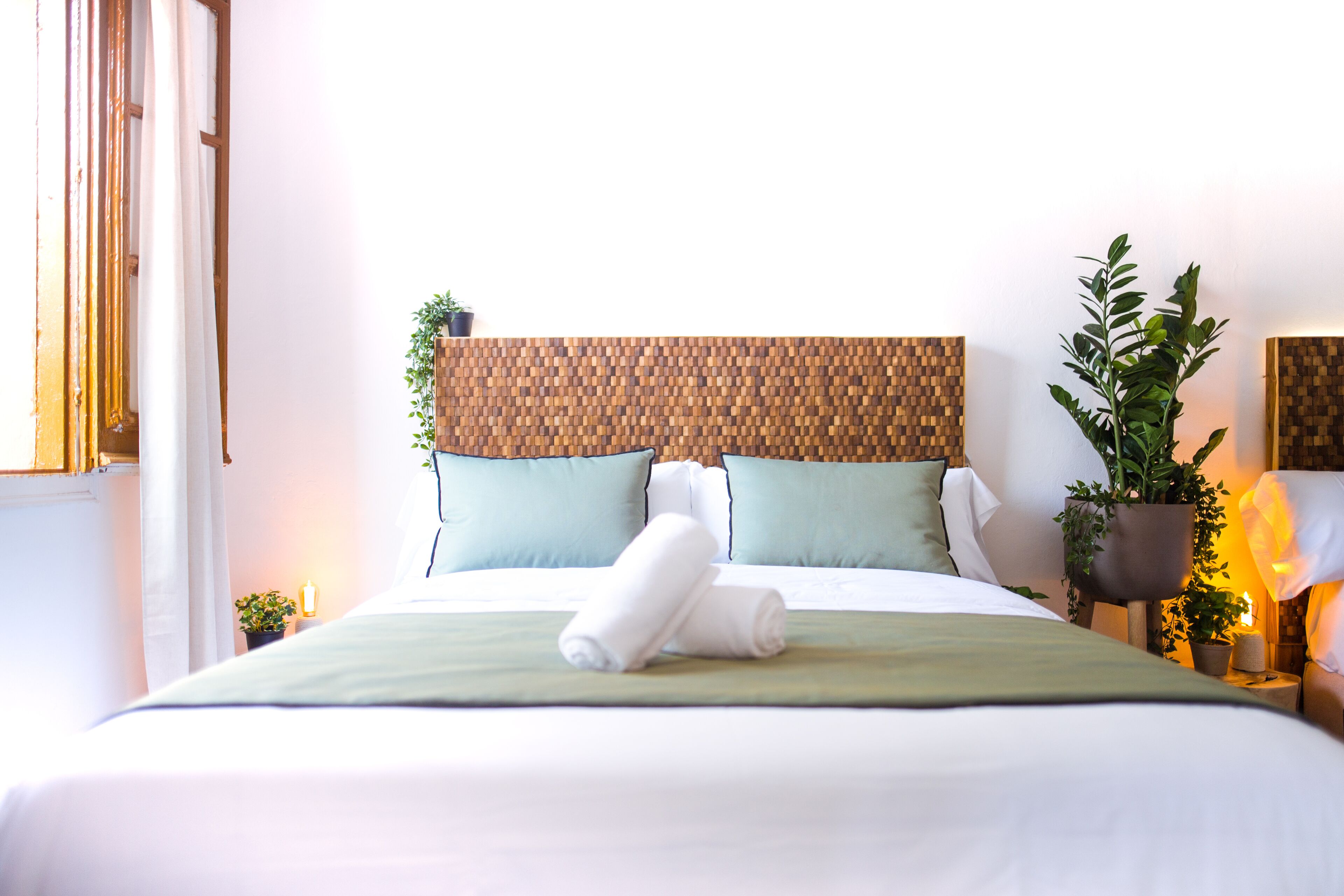 A neatly made bed with a green bedspread, two pillows, and a rolled towel. There is a wooden headboard, a nightstand with a lamp, and a potted plant.