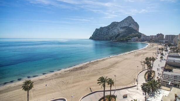 A coastal beach with a prominent rocky outcrop, a sandy shoreline, and a promenade lined with palm trees. - First line / Wifi / Spa / Seaviews / Heated pool / Pets (Calpe)