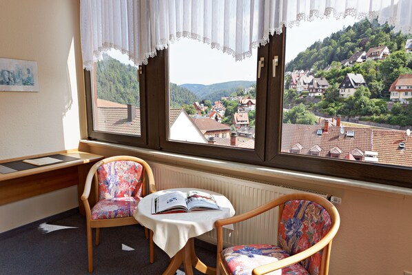 Quarto com vista para uma aldeia no topo de uma colina através de grandes janelas, uma mesa com um livro aberto e duas cadeiras.