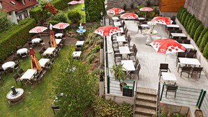 An outdoor patio with multiple tables and chairs, some with umbrellas, surrounded by greenery and a stone wall.