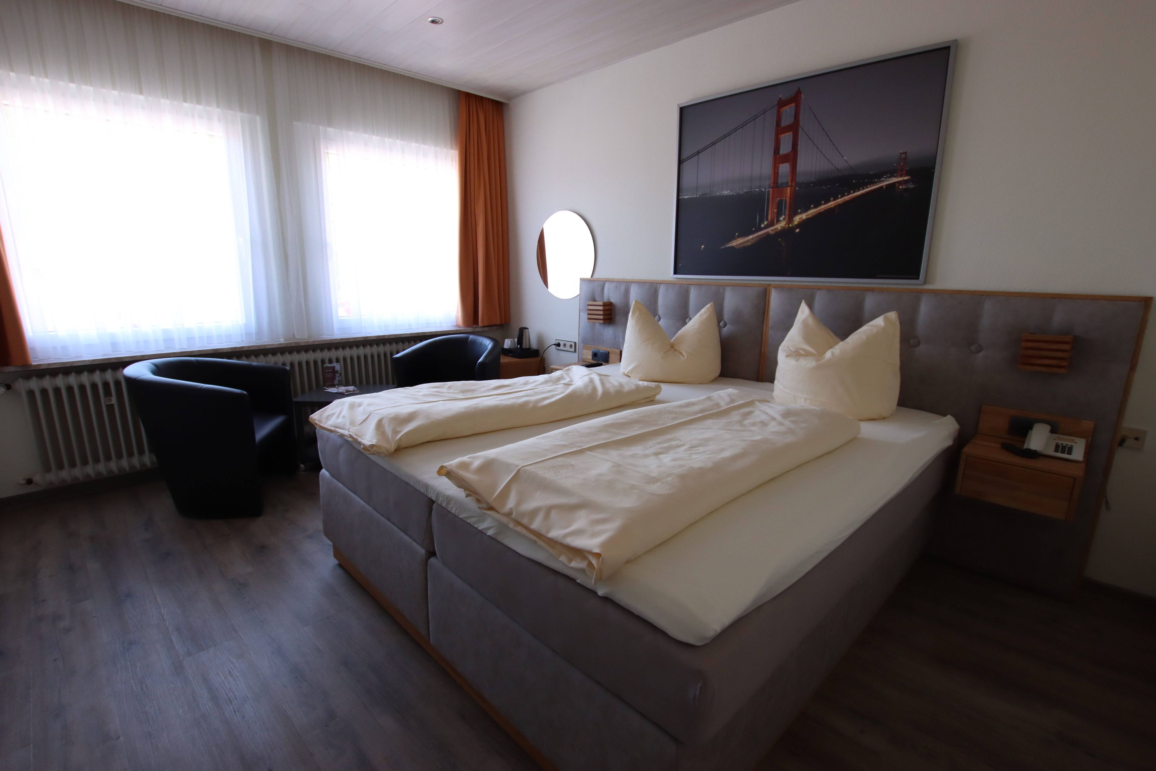 Ett hotellrum med en stor säng, ett skrivbord, en stol, ett fönster med gardiner och en tavla som föreställer en bro.