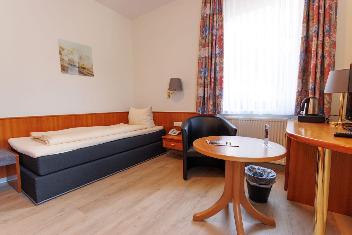 A hotel room with a sofa bed, a desk with a lamp, a chair, a trash bin, and a window with curtains.