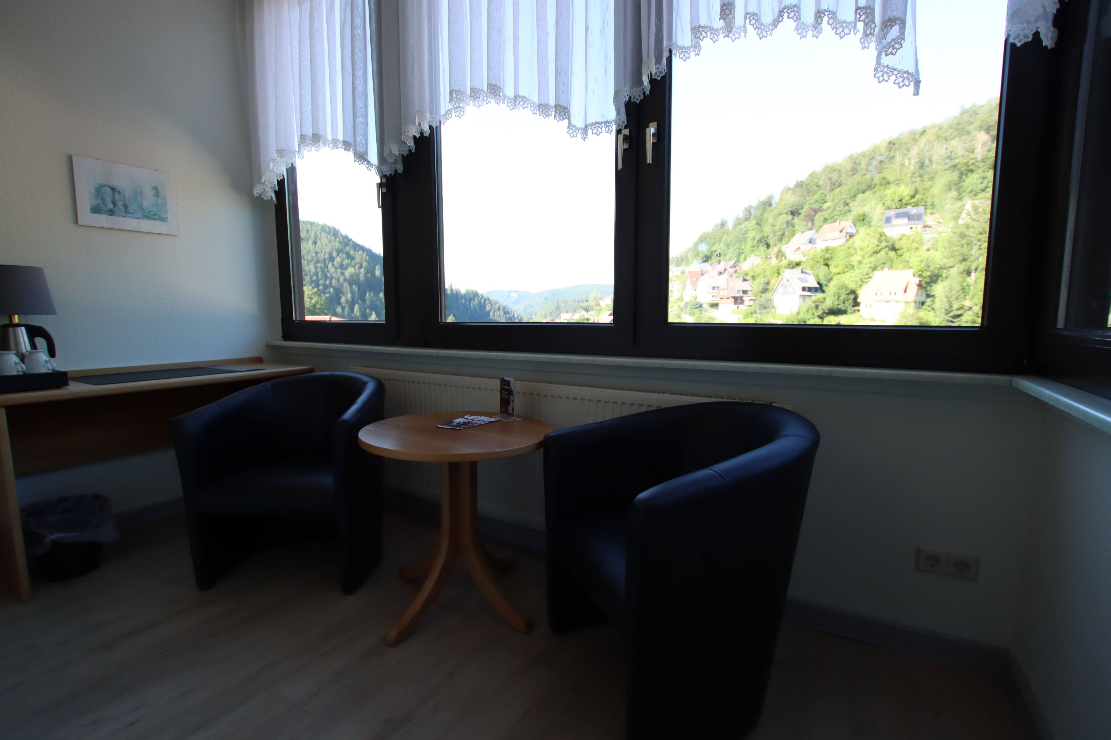 Una habitación con escritorio, dos sillas, una mesa redonda pequeña y ventana con vista a una colina.