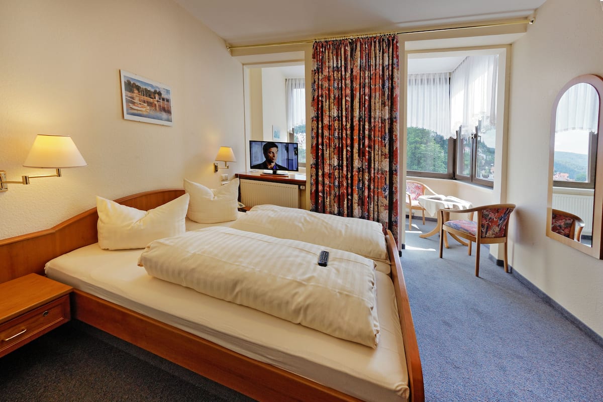 A hotel room with a double bed, two bedside tables, a TV, a balcony with a chair, and a view of the outdoors.