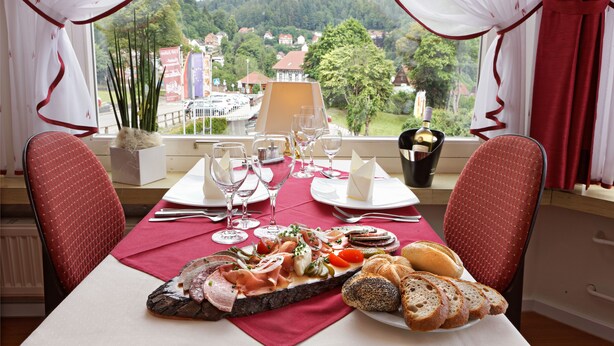 A dining table set with a variety of food and drinks, a view of a town through large windows, and red curtains.