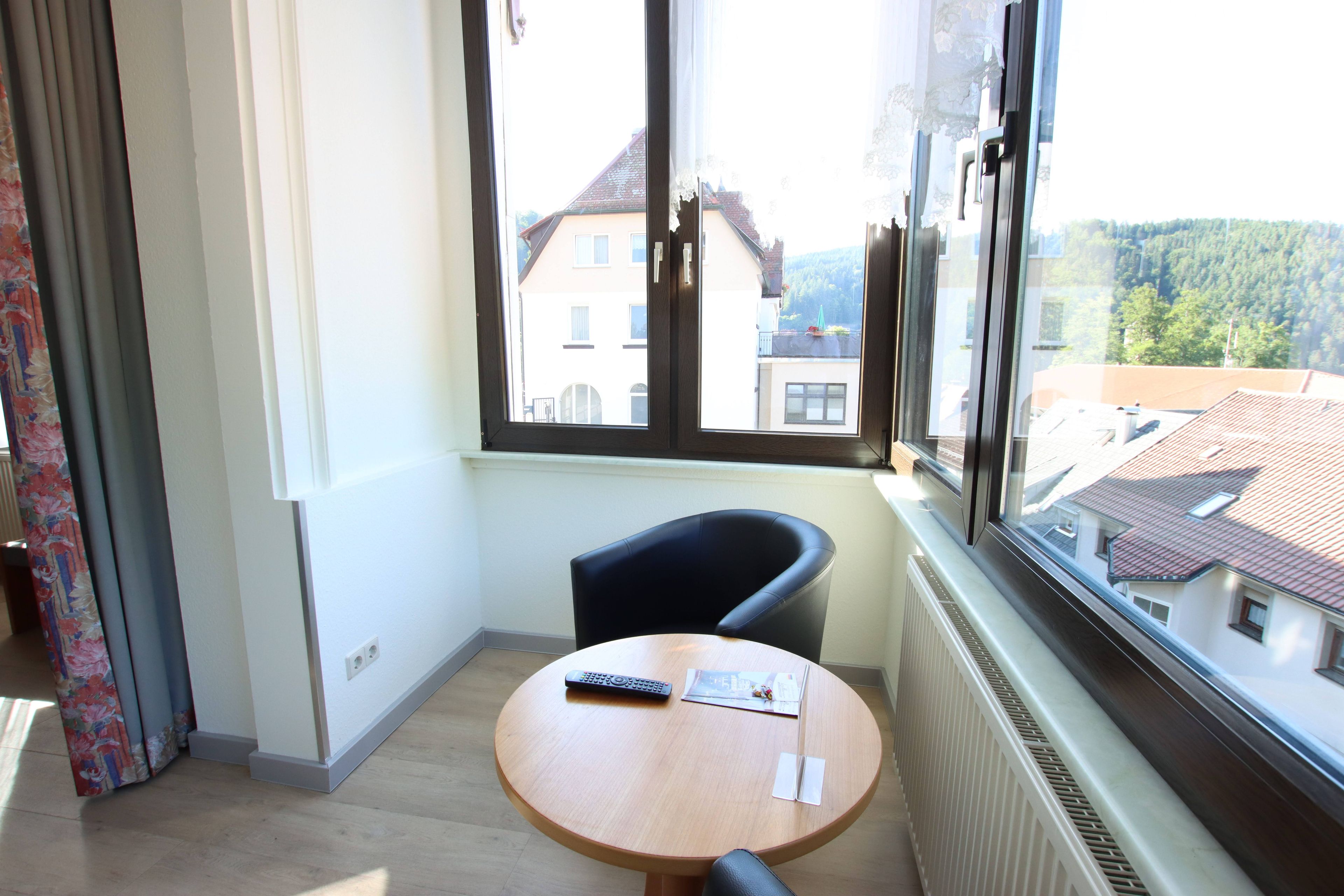 A room with a round table, a black chair, a window with a view of buildings and trees, and a radiator.