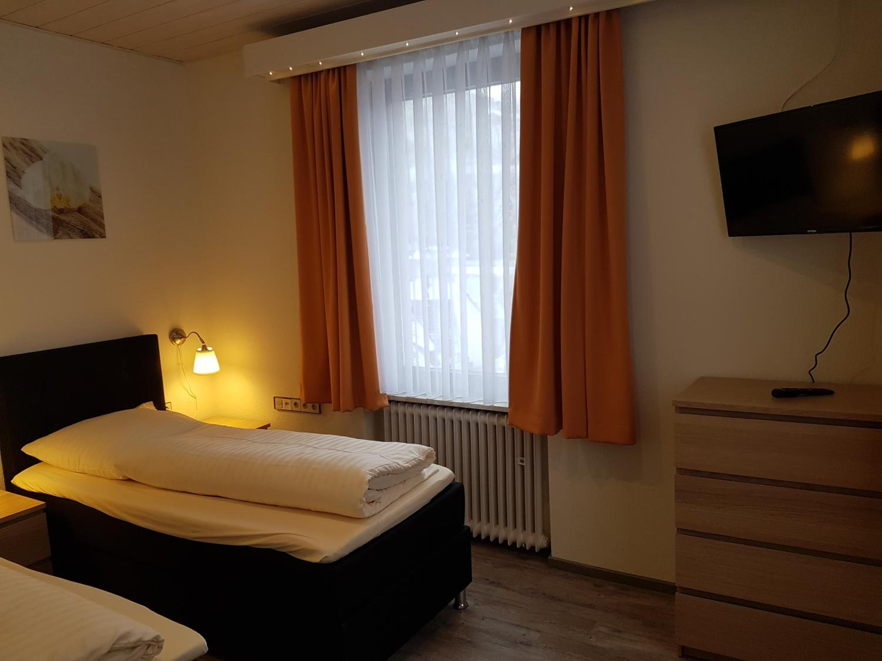 A hotel room with a bed, a nightstand with a lamp, a dresser, a television, and a window with curtains.