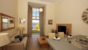 Ein modernes Wohnzimmer mit einer Couch, einem Couchtisch und einem Essbereich mit Tisch und Stühlen. Ein Kamin und ein Blick ins Freie sind ebenfalls vorhanden.