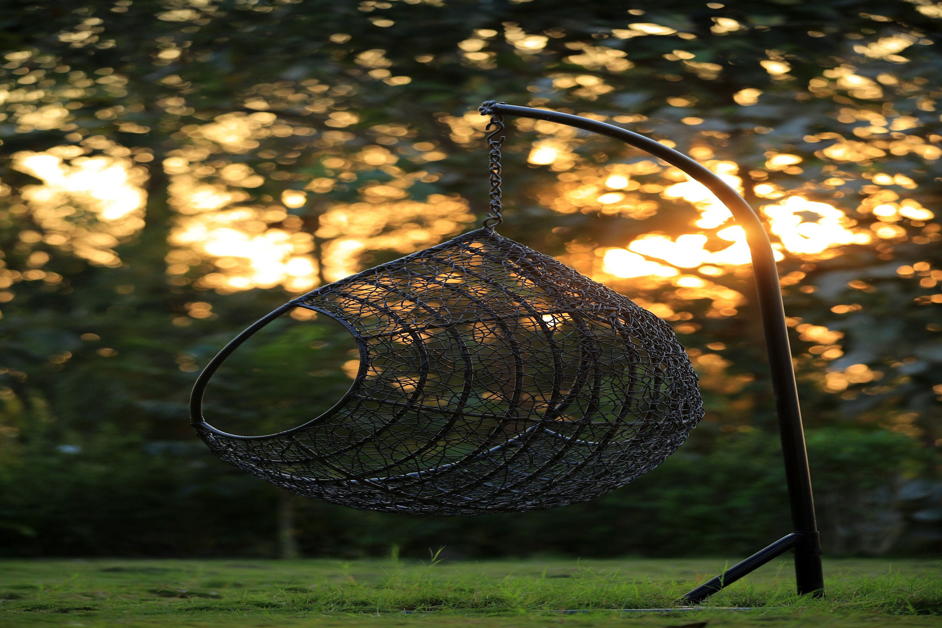 Un fauteuil suspendu de forme sphérique, fabriqué à partir d’un treillis métallique et soutenu par un pied en métal, placé dans un environnement extérieur avec des arbres et la lumière du soleil en arrière-plan.