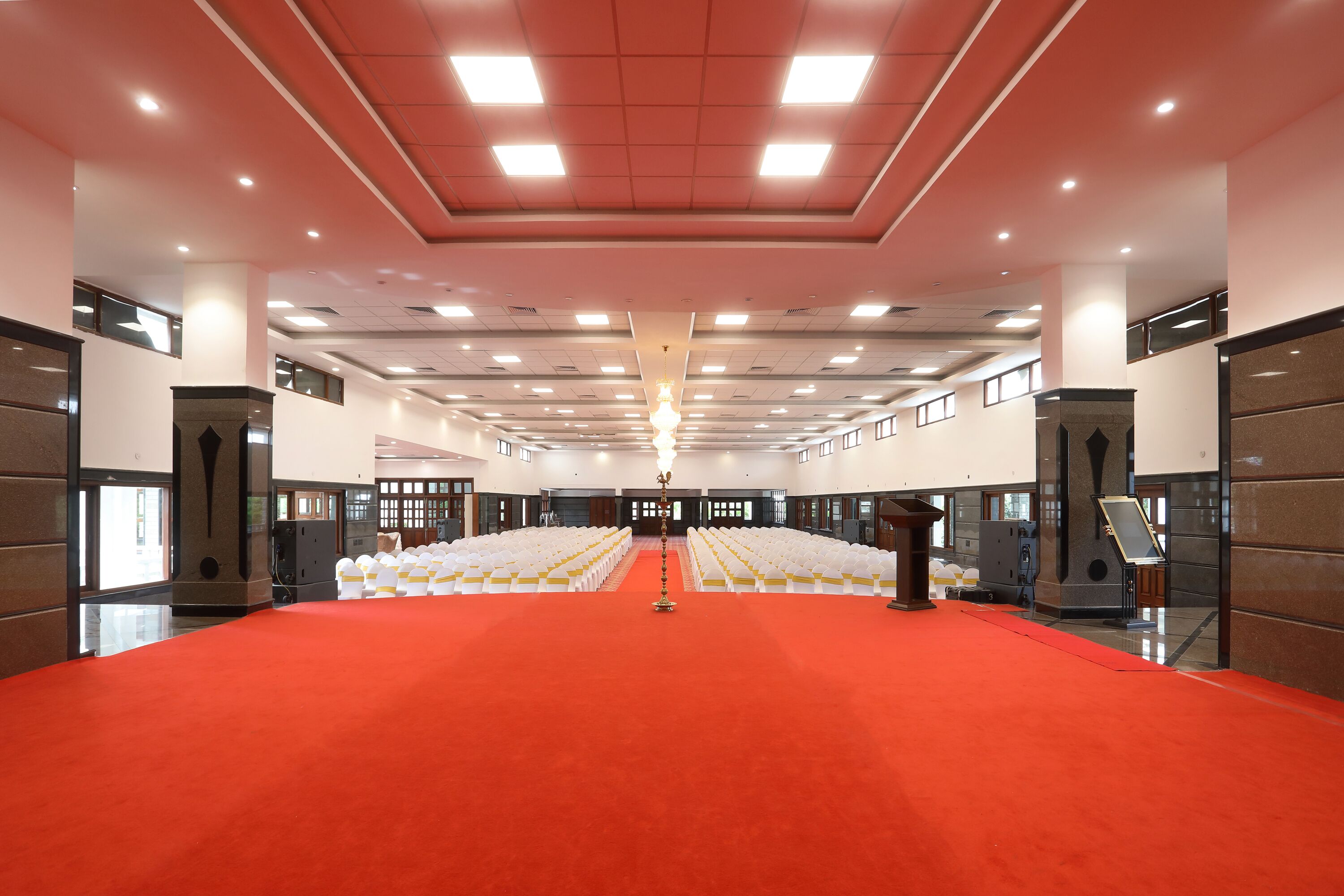 A spacious hall with a red carpet, rows of white chairs, and a podium at the front.