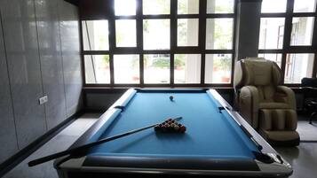 A pool table with balls, a pool cue, and a massage chair in a room with large windows.