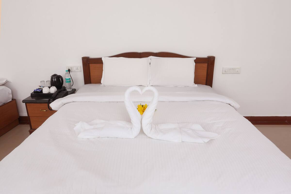 A neatly made bed with a heart-shaped towel arrangement, a bedside table with a water bottle, a phone, and a cup.