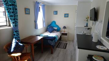 Une pièce de taille modeste comprenant un lit, un petit bureau, une chaise, un téléviseur et un coin cuisine.