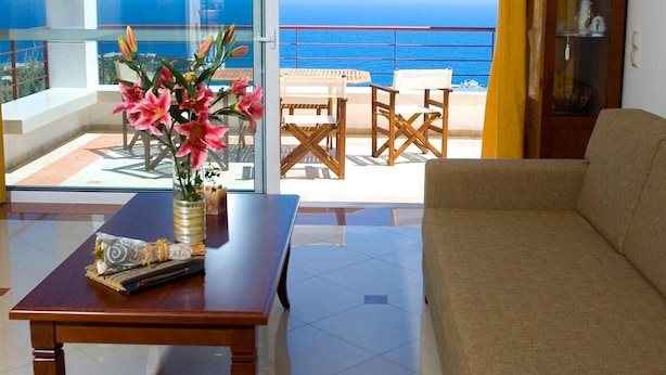 Een woonkamer met een bank, een houten salontafel en een bloemenvaas. Er is een balkon met uitzicht op de zee en een zithoek met houten stoelen en tafels.