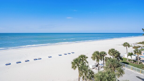 Une plage de sable blanc, avec une eau d’un bleu limpide et des palmiers. Des chaises de plage et des parasols sont alignés le long du rivage.
