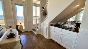 Une cuisine avec des meubles blancs, un plancher en bois et une vue sur l’océan par une porte.