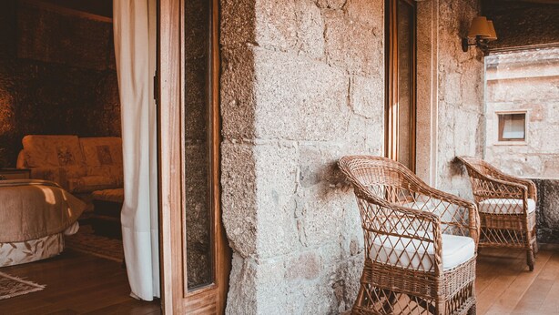 Una habitación con pared de piedra, sillas de mimbre, suelo de madera y una cama con colcha blanca.