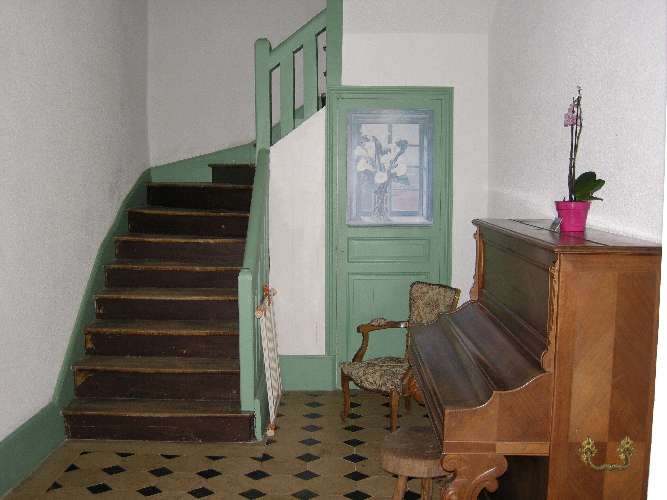 A staircase with green railings, a door with a floral window, a chair, and a wooden cabinet with a plant.