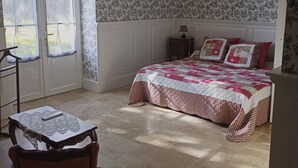 A bedroom with a large window, a bed with a patterned quilt, a small table with a lace tablecloth, and a chair.