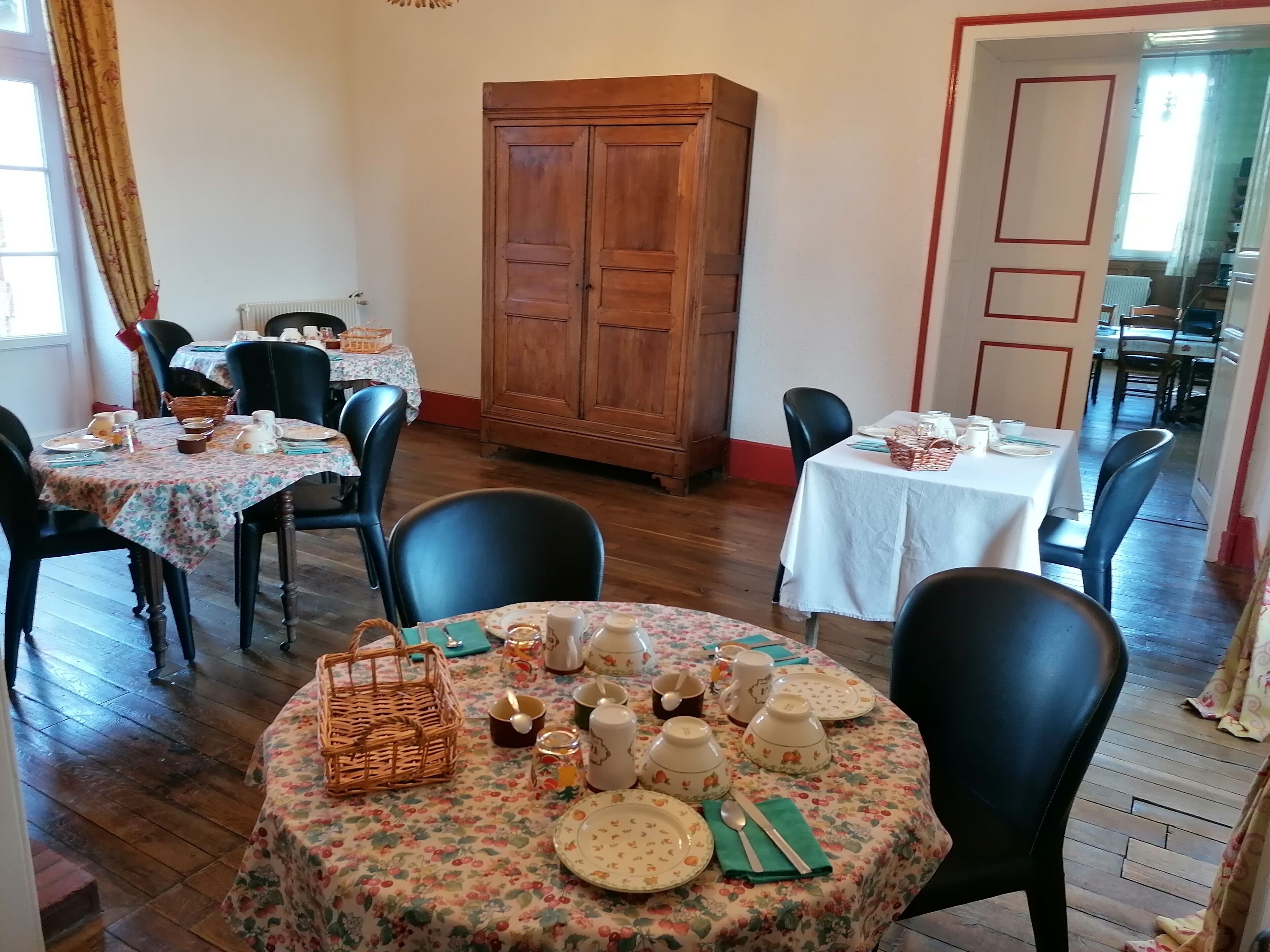 Un comedor con mesas redondas dispuestas para comidas, armarios de madera y vista a la cocina por una puerta abierta.