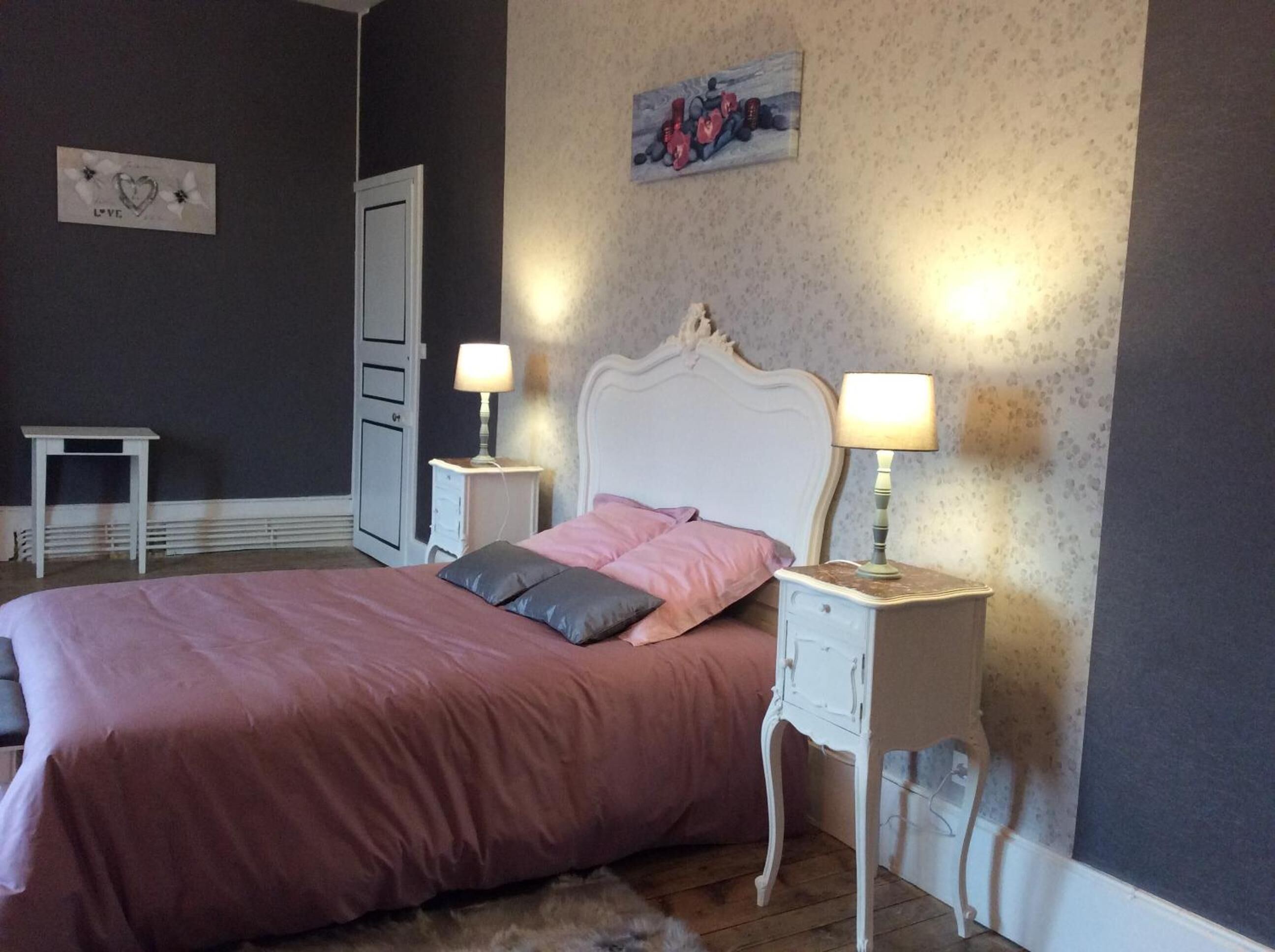 Une chambre à coucher avec un lit, deux tables de chevet avec des lampes, une commode, une porte et un mur orné d’un motif floral.