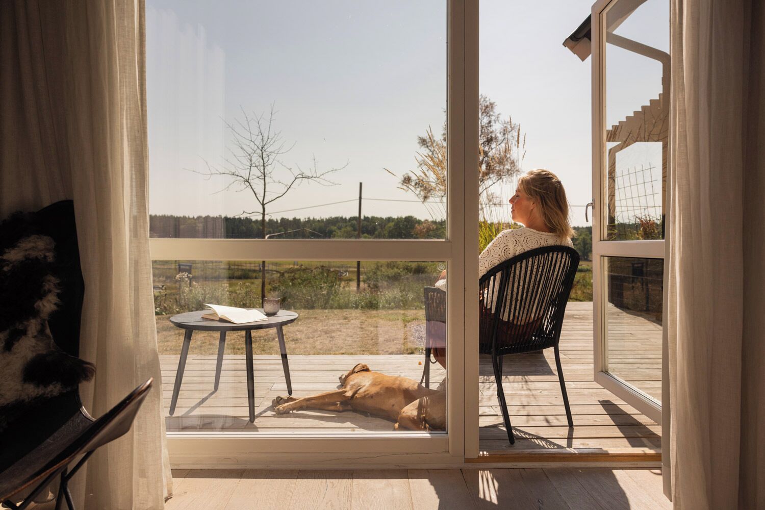 A woman sitting on a chair by a glass door, looking out at a dog lying on the deck.