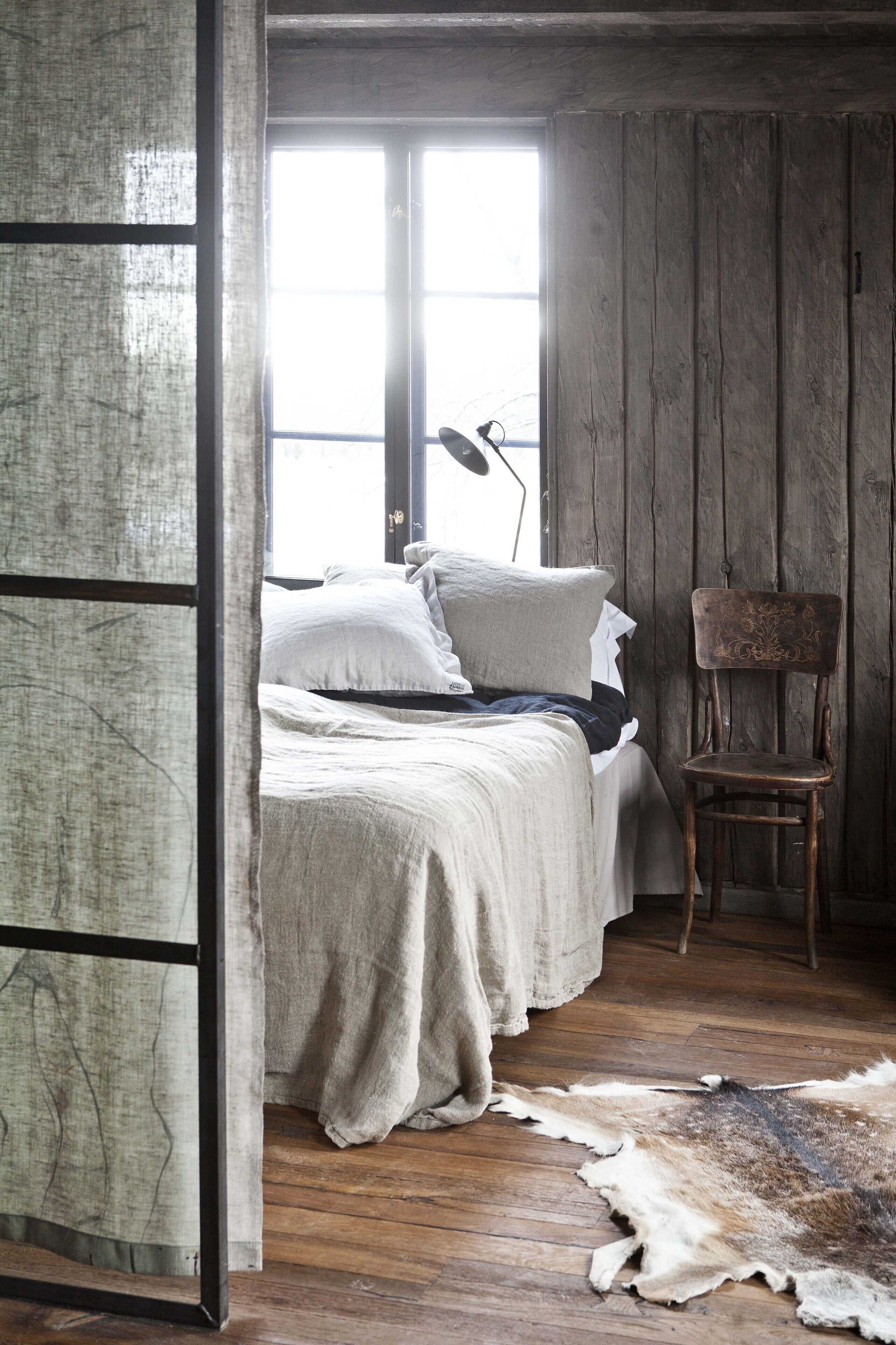 Un dormitorio rústico con piso de madera, una cama con ropa de cama blanca, una silla y un ventanal grande.