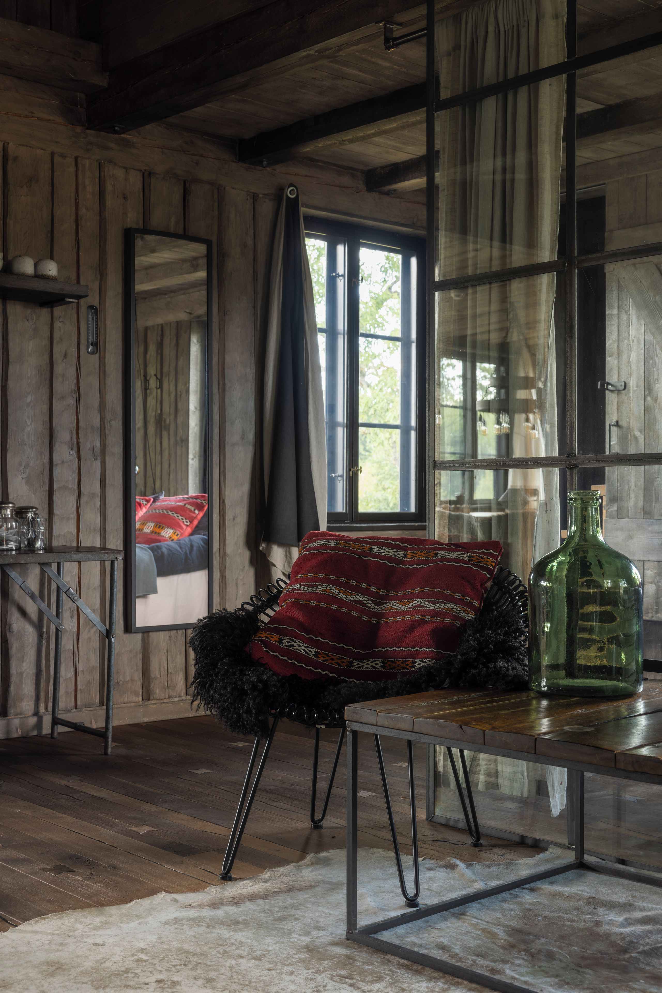 Une pièce rustique avec un plancher en bois, une table à ossature métallique, un coussin rouge à motifs, une bouteille verte et une fenêtre avec des rideaux.