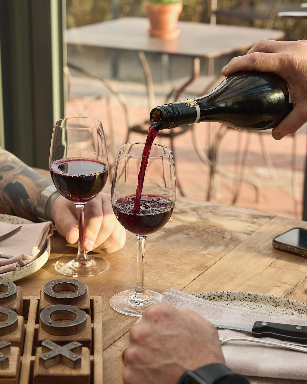 Una persona sirve vino tinto en una copa sobre una mesa de madera, con un cuchillo y una tabla de cortar cerca.