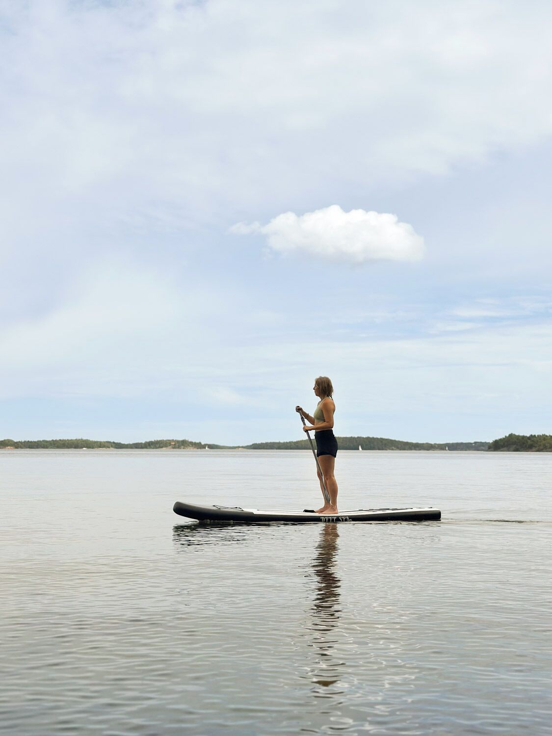 A person standing on a paddleboard in a calm body of water with a distant shoreline and a cloudy sky.