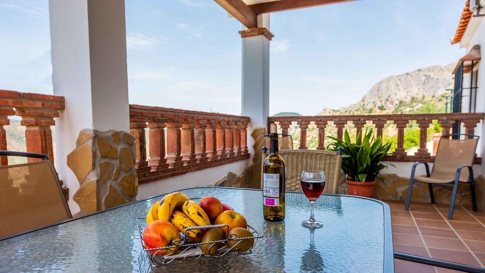 A glass table with a bottle of wine, a glass of red wine, and a bowl of fruit on a balcony with a view of mountains.
