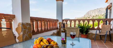 A glass table with a bottle of wine, a glass of red wine, and a bowl of fruit on a balcony with a view of mountains.