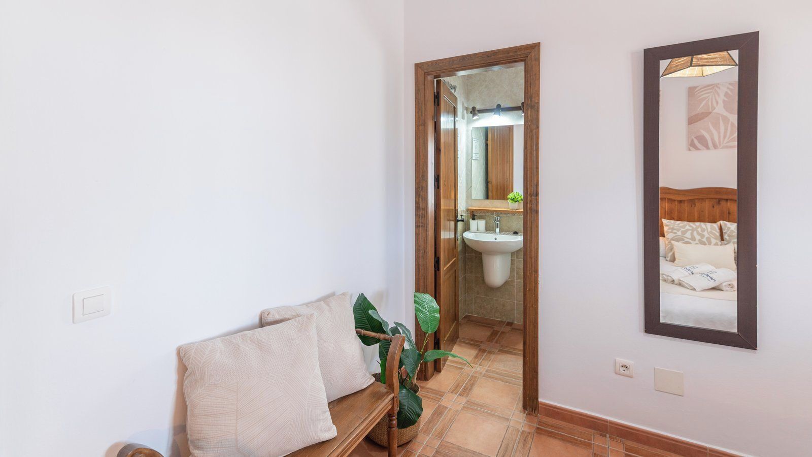 Une chambre d’hôtel comprenant un lit, un petit coin salon avec des coussins, une plante en pot, une salle de bain avec un lavabo et un miroir, et un tableau encadré accroché au mur.