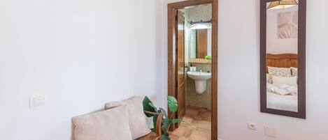 Une chambre d’hôtel comprenant un lit, un petit coin salon avec des coussins, une plante en pot, une salle de bain avec un lavabo et un miroir, et un tableau encadré accroché au mur.