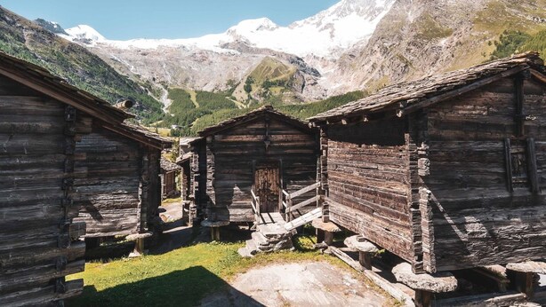 Ein rustikales Holzhaus mit einer Steintreppe, die zu einer Holztür führt, vor einer Kulisse aus schneebedeckten Bergen und blauem Himmel.