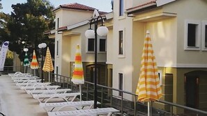 Fila de tumbonas blancas con sombrillas a rayas, zona de piscina y un edificio con tejado de baldosas.