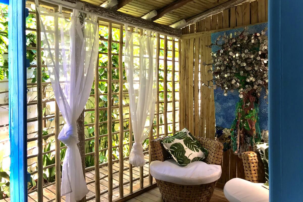 A room with a bamboo lattice window, a wicker chair with a cushion, and a decorative wall with a tree and hanging ornaments.