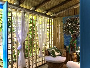 A room with a bamboo lattice window, a wicker chair with a cushion, and a decorative wall with a tree and hanging ornaments. - Casa da Gloria (Porto Seguro)