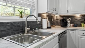 A modern kitchen with white cabinets, a stone backsplash, a stainless steel sink, and a coffee maker.